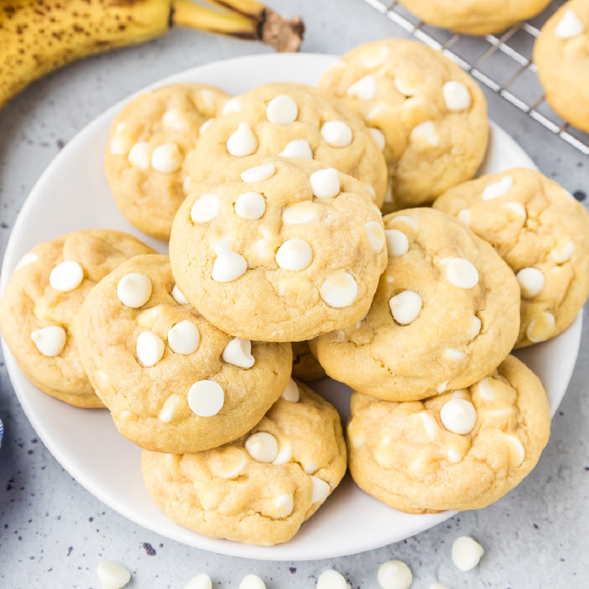 Square view of a plate of banana pudding cookies topped with white chocolate chips on a counter with a banana and a wire cooling rack in the background.