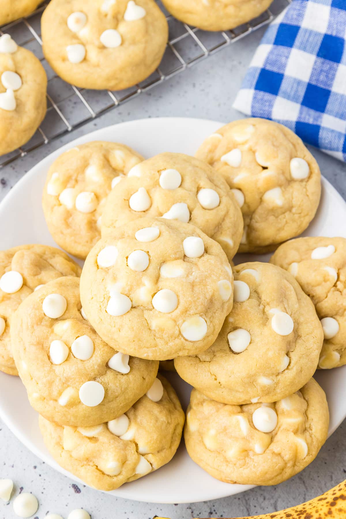 A full plate of banana pudding cookies topped with white chocolate chips from above on a counter next to a wire cooling rack with more cookies.