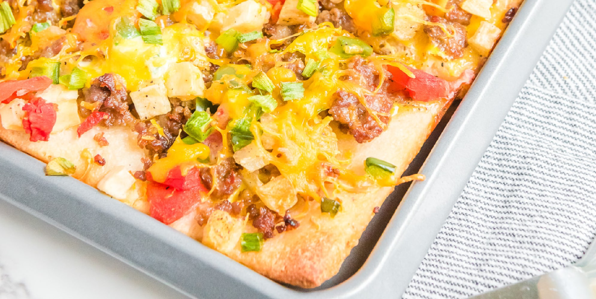 Wide view of a baking sheet full of breakfast pizza topped with eggs, sausage, cheese and vegetables turned at an angle to see the corner of the pan.