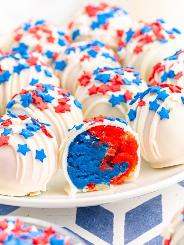 A plate of cake balls dipped in white chocolate and coated in red and blue star sprinkles with one cake ball cut open to reveal red and blue cake inside.