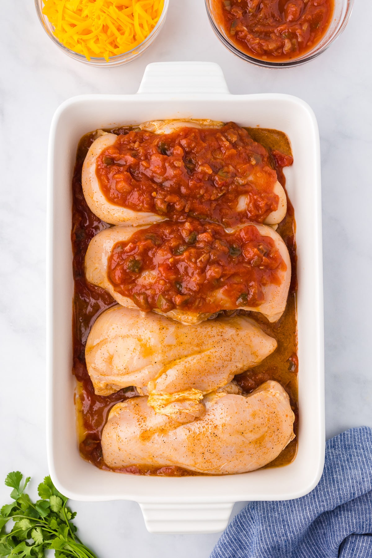 Four raw chicken breast in a rectangular casserole dish with salsa on the bottom of the pan and with two chicken pieces topped with salsa.