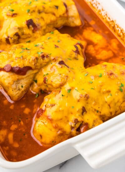 Chicken breasts in a tomato sauce covered in cheddar cheese in a pan at an angle from the side.