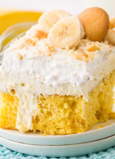 Close up side view of a slice of banana pudding cake topped with layers of pudding, whipped cream, sliced banana and vanilla cookies on a plate from the side.