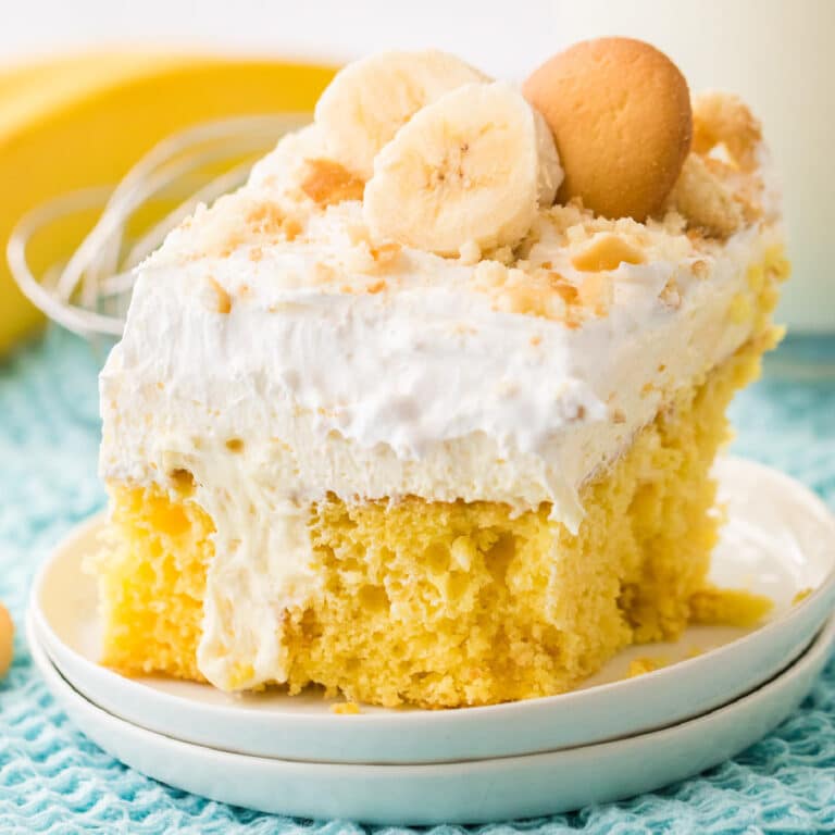 Close up side view of a slice of banana pudding cake topped with layers of pudding, whipped cream, sliced banana and vanilla cookies on a plate from the side.