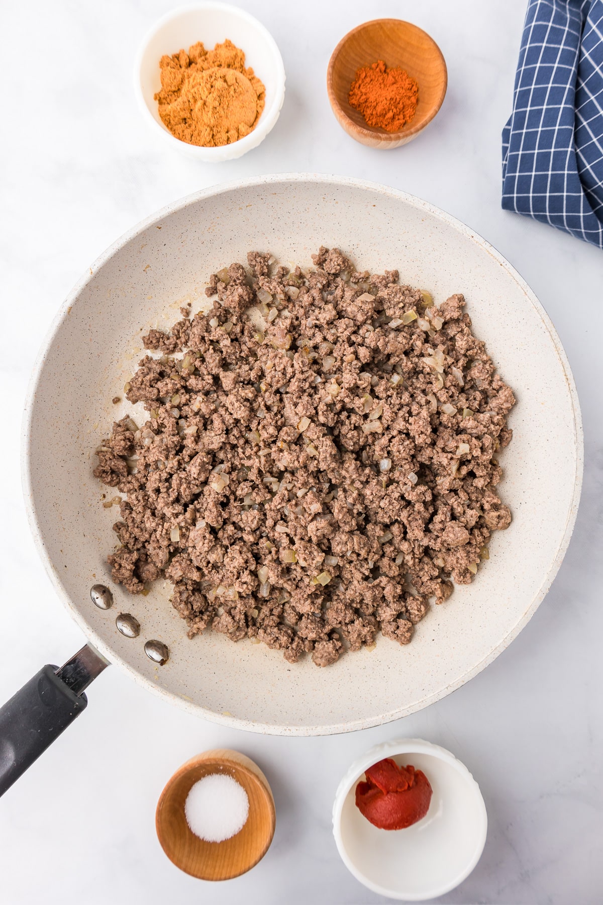 Browned ground beef in a skillet from overhead with seasonings and tomato paste in small bowls nearby on the counter.