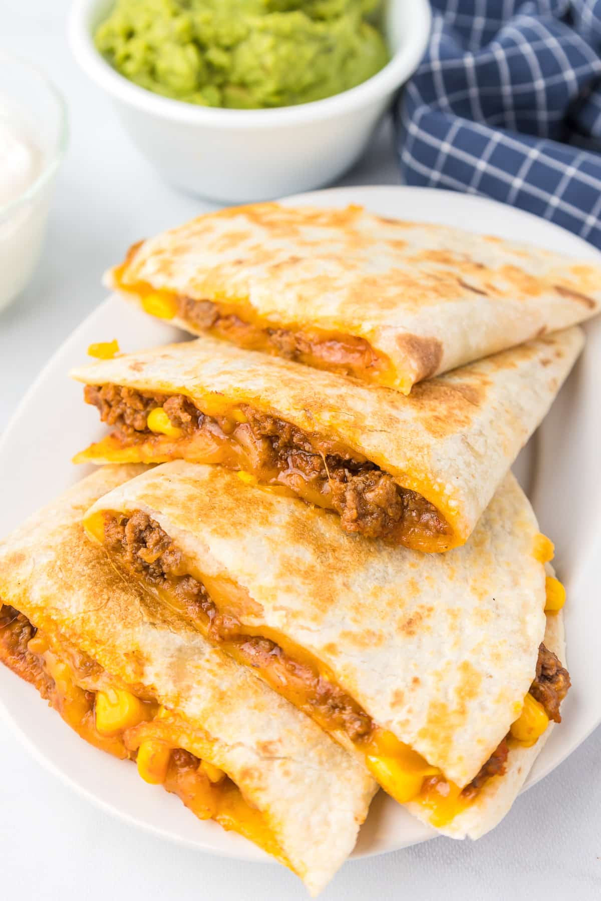 Four ground beef, cheese and corn quesadillas laid in a row on a plater from the side with a bowl of guacamole in the background.