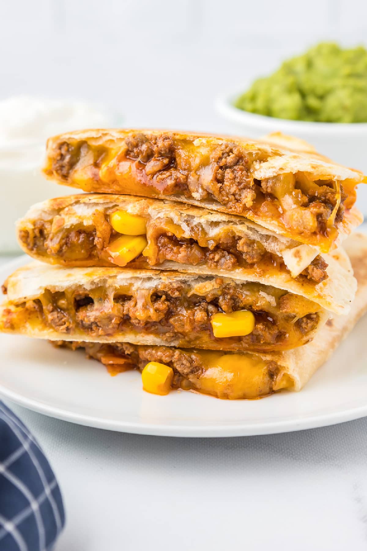 Side view of a beefy cheesy quesadilla stacked four high on a plate with piece of corn in the filling and guacamole in a bowl in the background.