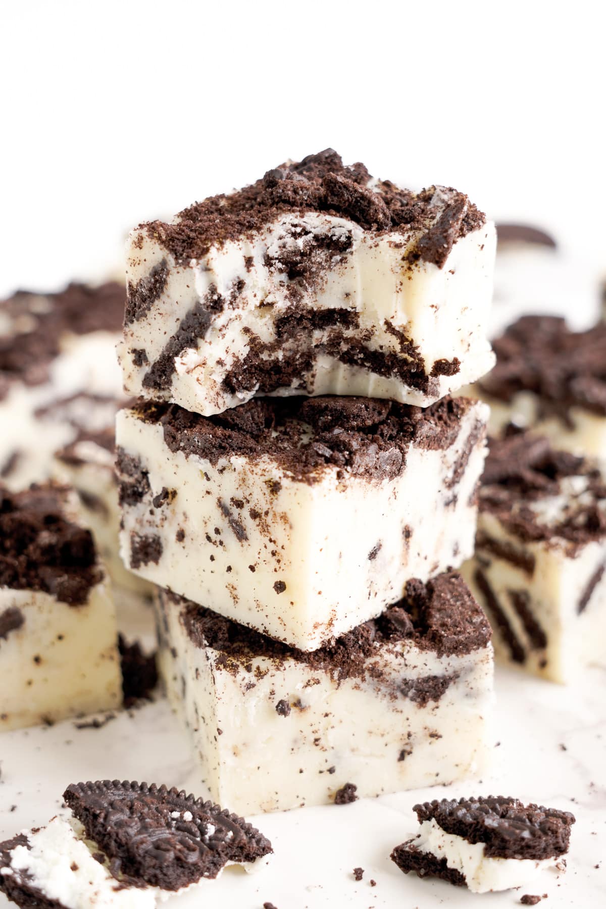 Three pieces of cookies and cream fudge stacked on top of each other with the top piece missing a bite from the side, and more fudge nearby on the counter.
