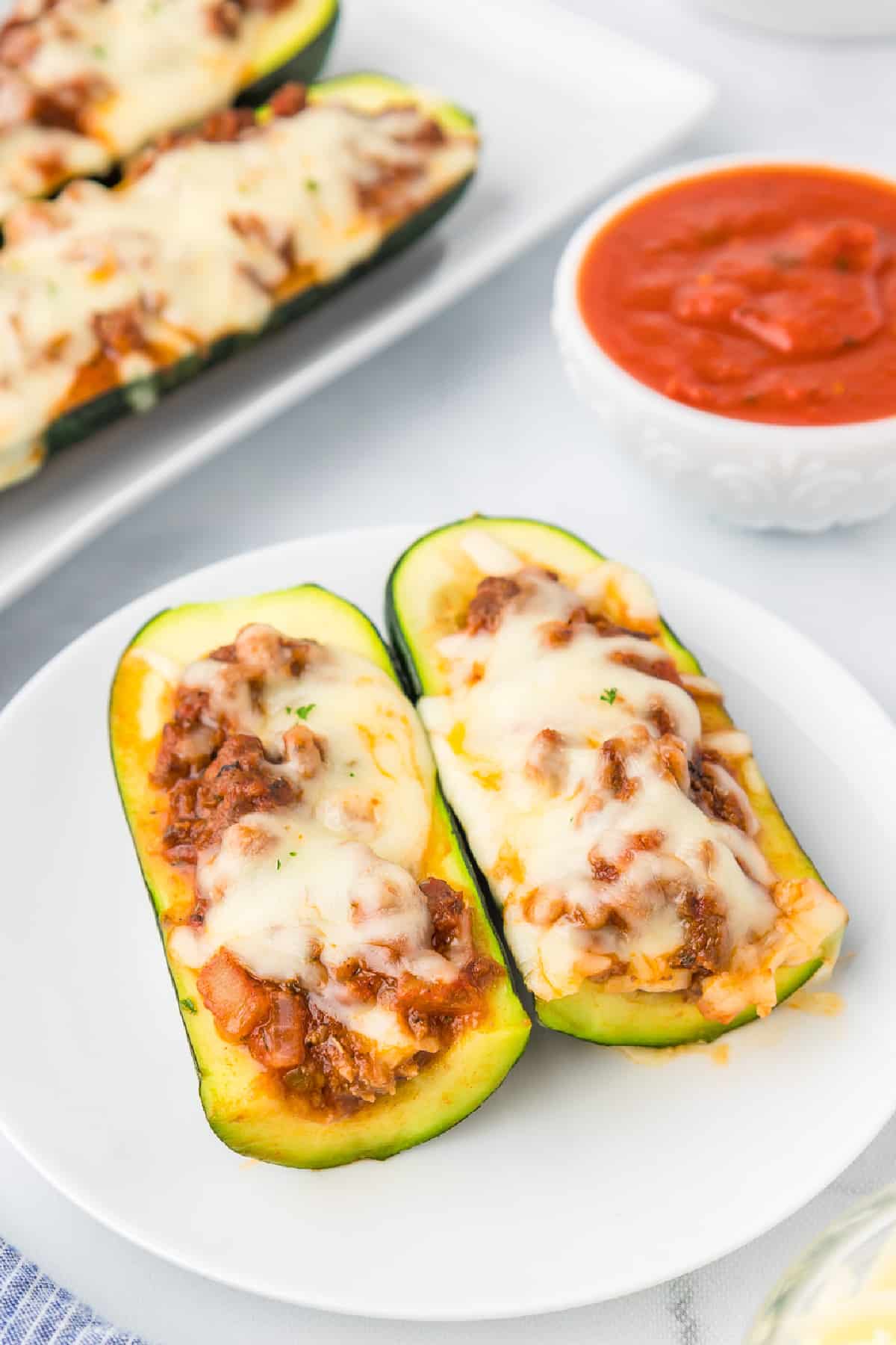 A stuffed zucchini boat sliced in half on a plate with marinara in a bowl nearby and more zucchini on a platter in the background on the counter.