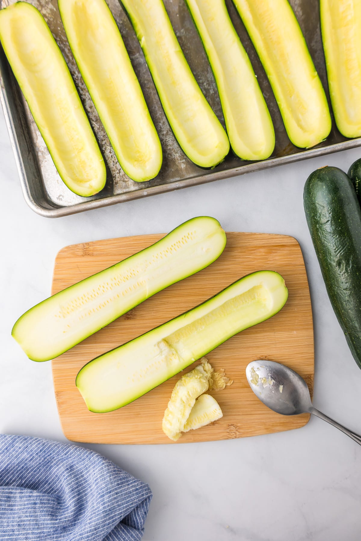 Two slices of zucchini sliced longways being scooped with a spoon on a cutting board with more hollowed out zucchini lines up on a baking sheet nearby.