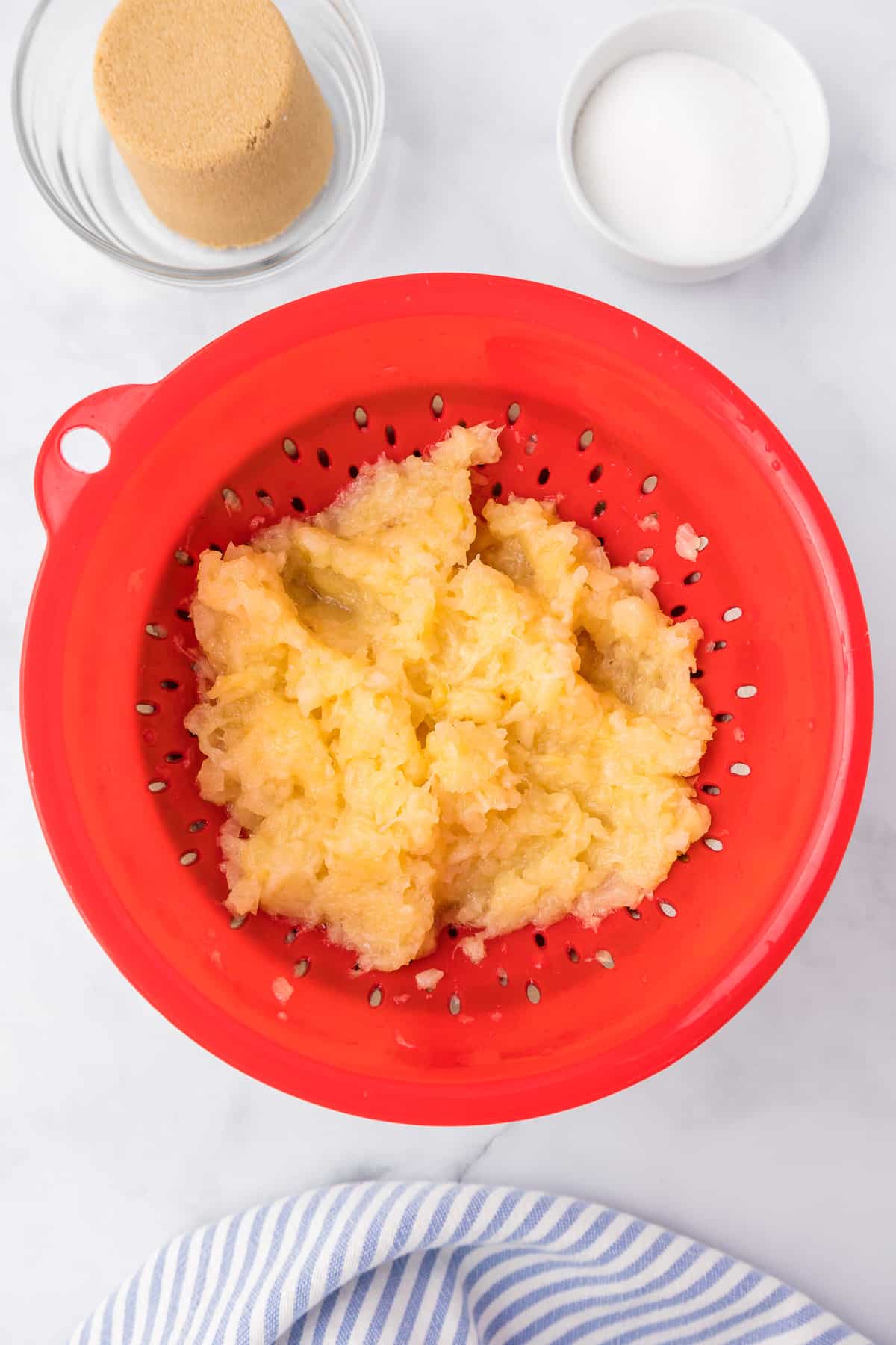 Pineapple being drained in a colander from above with brown sugar and sugar nearby in bowls.