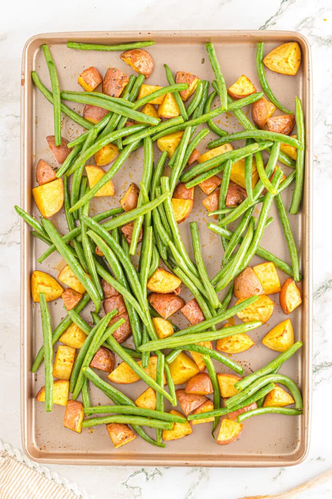 Green beans being added to a sheetpan of partially roasted potato cubes from above.