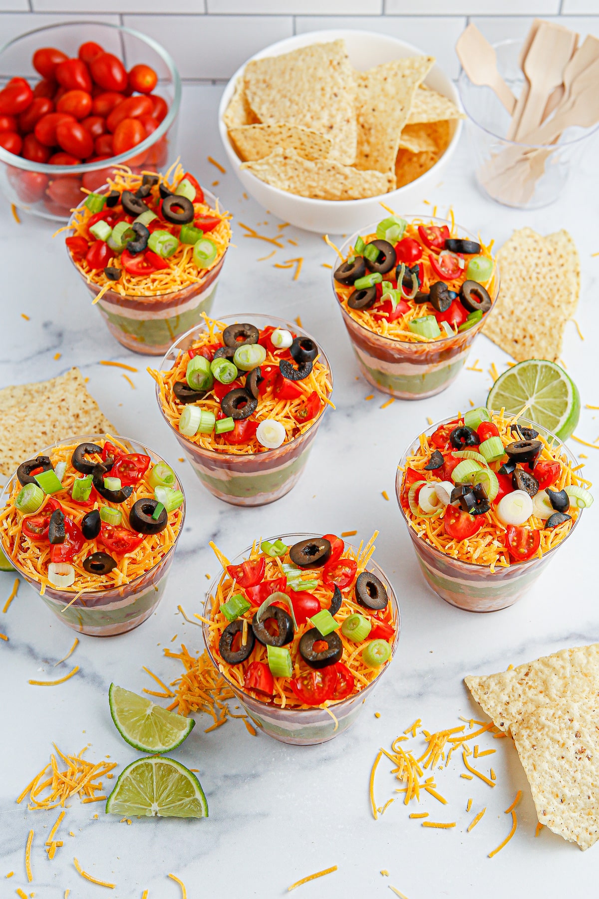Six seven layer dip cups from overhead on a counter with more toppings, chips and lime slices nearby on the counter.