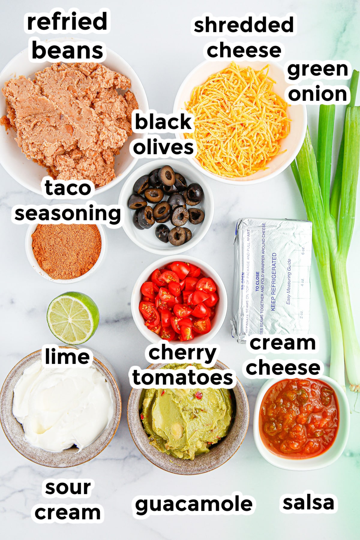 Ingredients for seven layer dip cups on a counter in bowls from overhead with text labels.