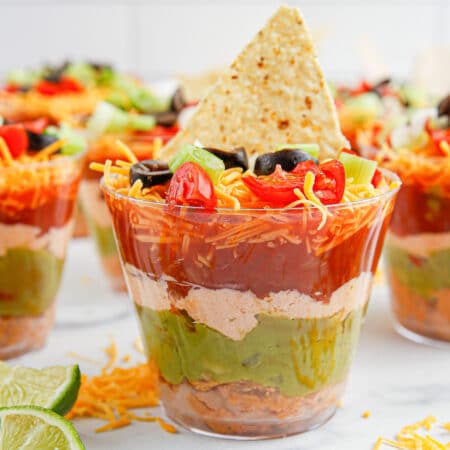 Square up close view of seven layer dip cups focused in one one cup to see the layers topped with a chip with more cups in the background.