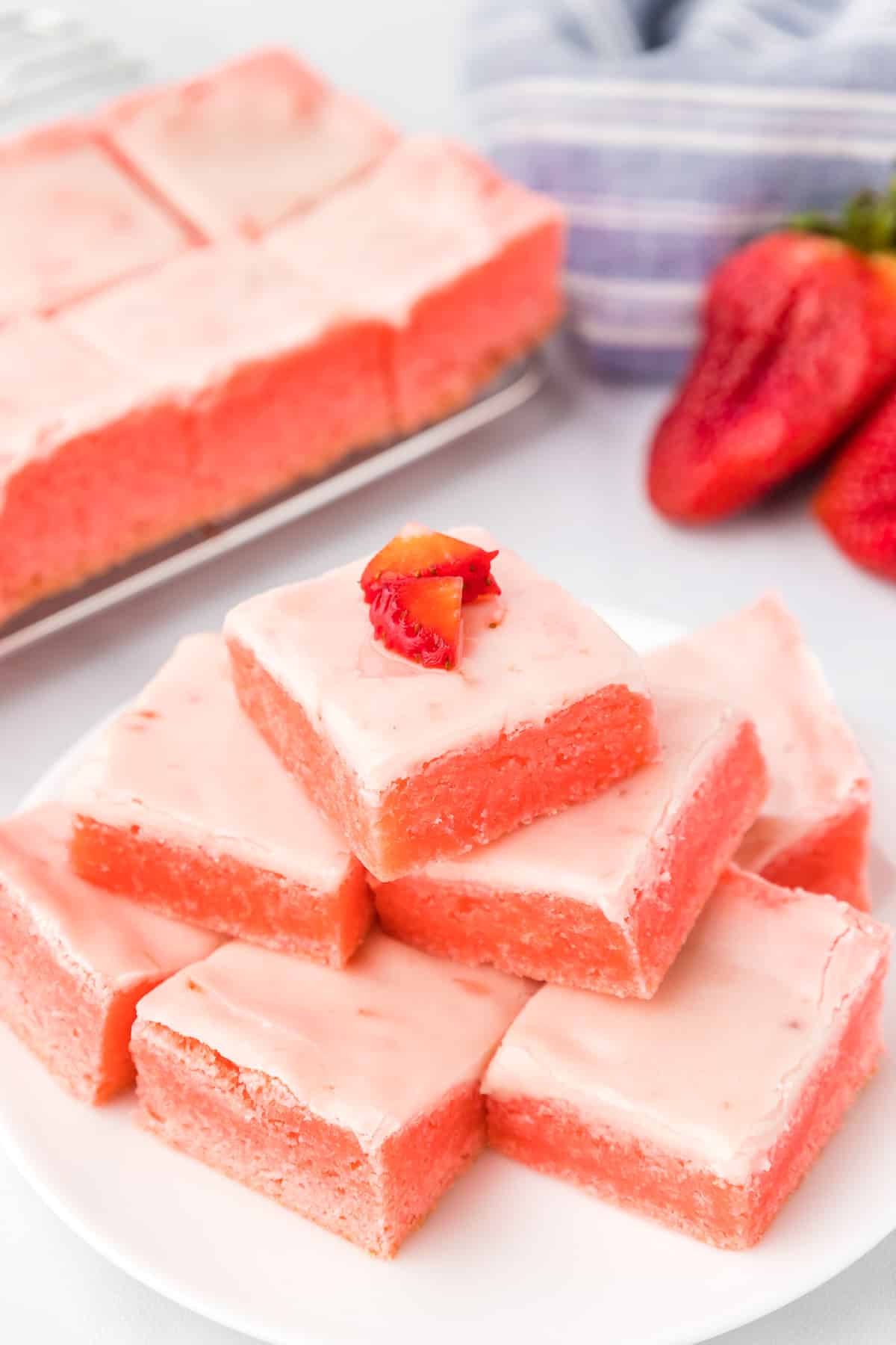 Strawberry brownies with glaze stacked on a platter with the top piece with fresh strawberry pieces on top and more strawberry brownies and fresh strawberries on a plater in the background on the counter.