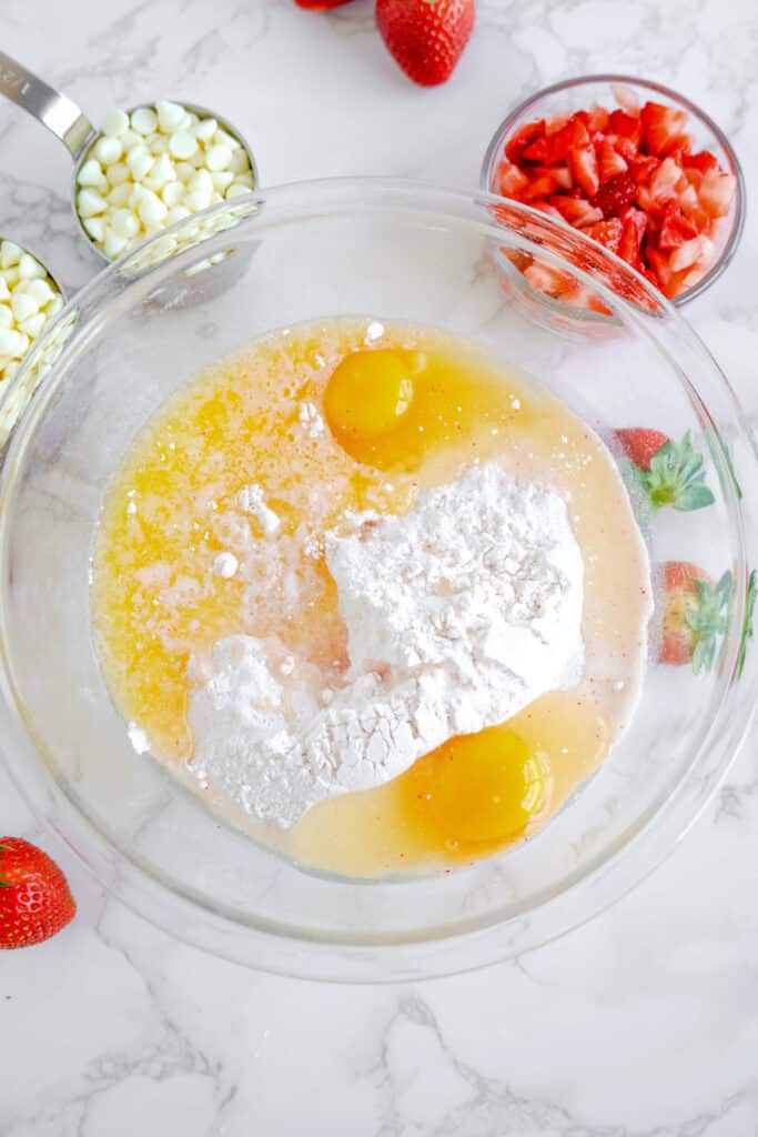 Eggs, cake mix and melted butter added together in a large mixing bowl from overhead with chopped strawberries and white chocolate chips nearby in bowls on the counter.