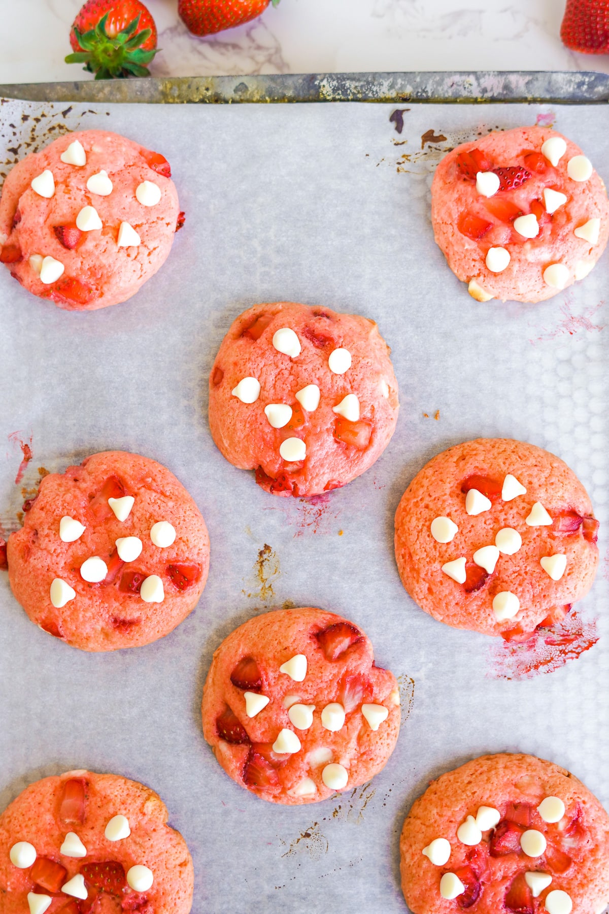 Strawberry cookies cooling on a baking sheet being topped with extra white chocolate chips and strawberries.