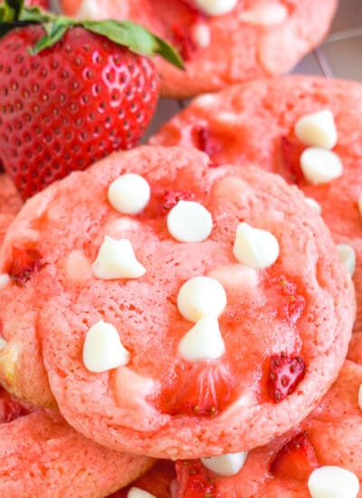 Pink strawberry cookies with white chocolate chips piled together but close up on the top cookie. A few fresh strawberries are next to the cookies.