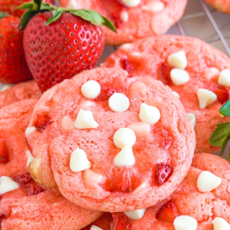 Pink strawberry cookies with white chocolate chips piled together but close up on the top cookie. A few fresh strawberries are next to the cookies.