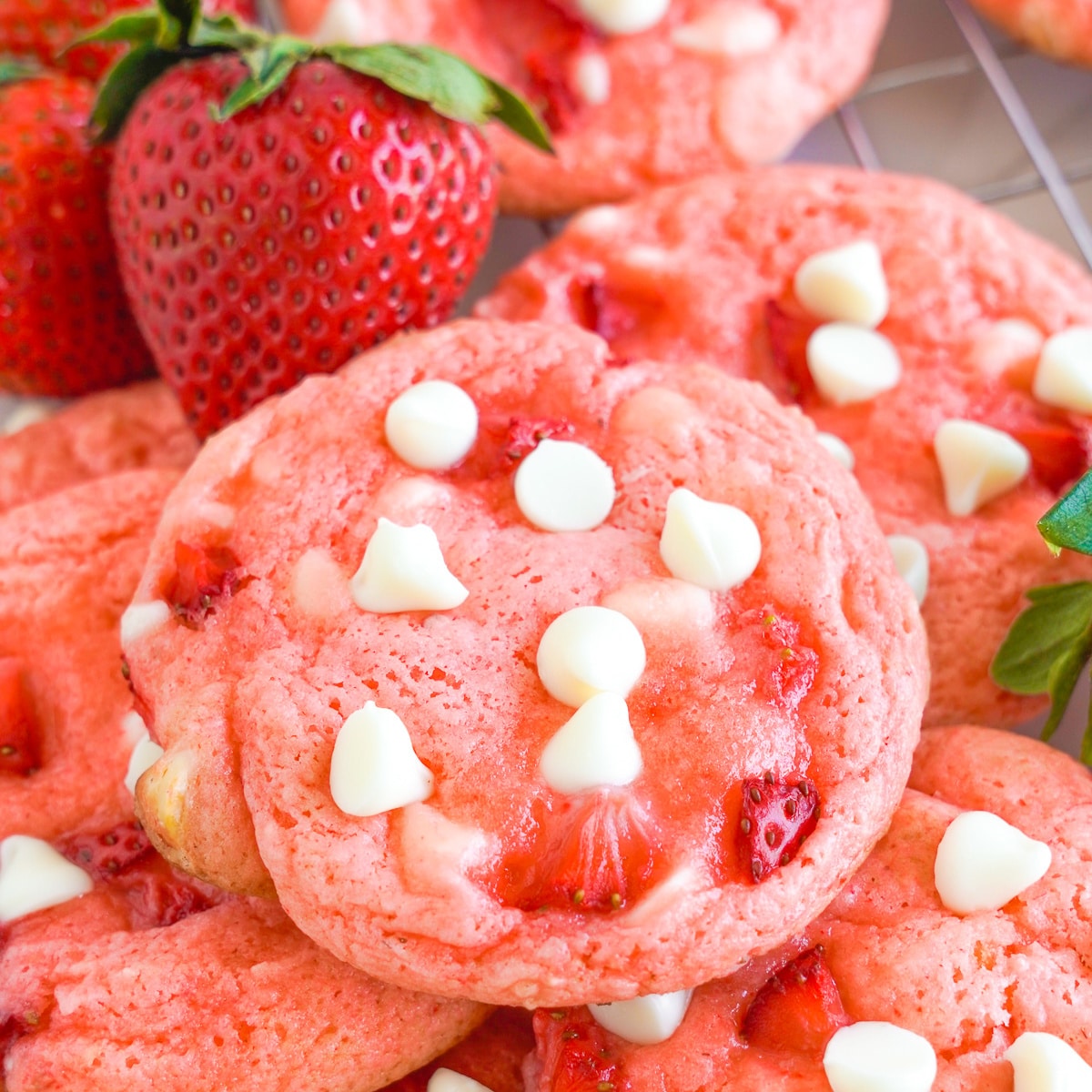 Pink strawberry cookies with white chocolate chips piled together but close up on the top cookie. A few fresh strawberries are next to the cookies.