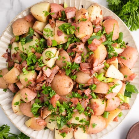 Square close up view of a platter full of cooked red potatoes, bacon pieces, green onions and parsley from above.