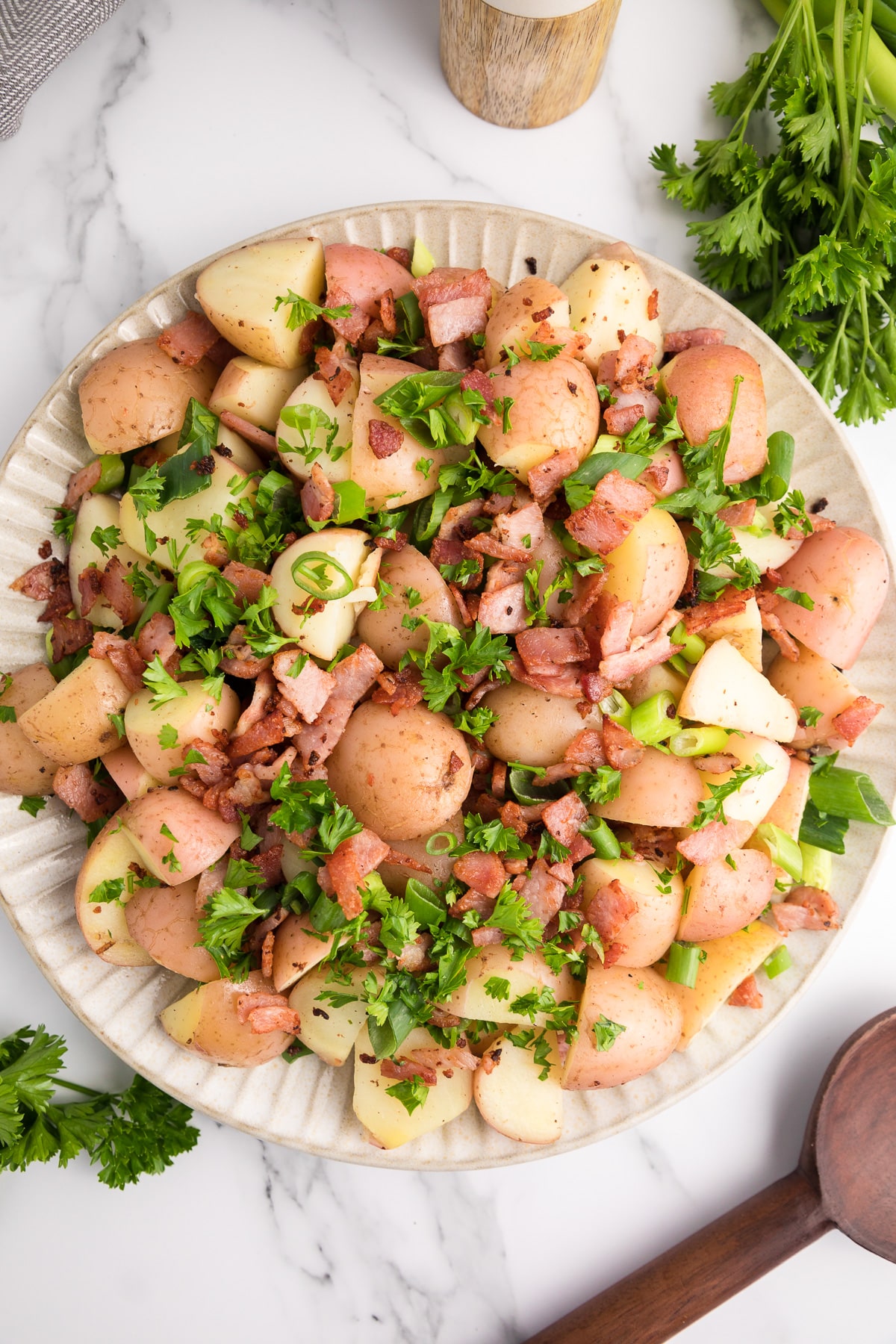Warm german potato salad on a large platter topped with bacon pieces, green onion and parsley on a counter from above.