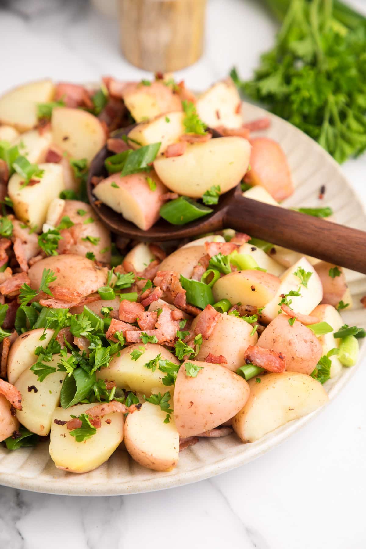 Close up of a platter full of warm german potato salad being scooped with a spoon topped with bacon, parsley and green onion pieces.