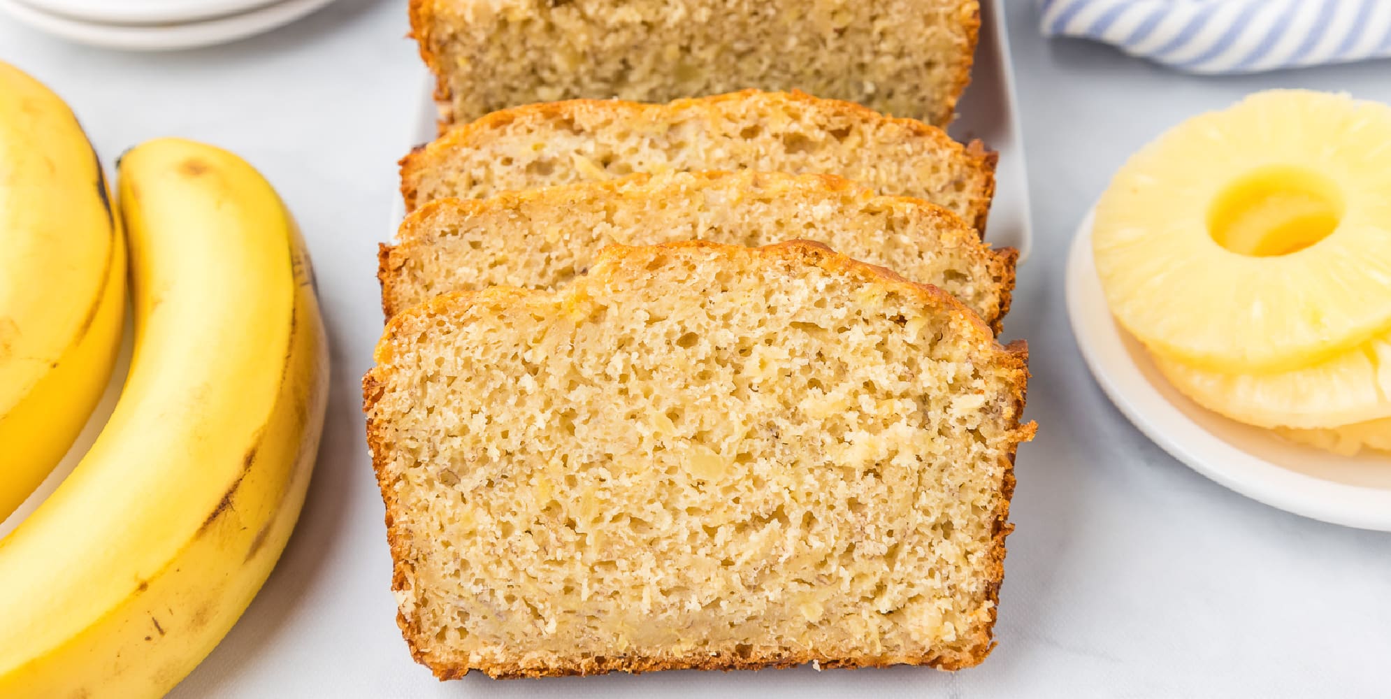 Slices of pineapple banana bread leaning back onto a platter close up with pineapple rings and bananas on the counter nearby.