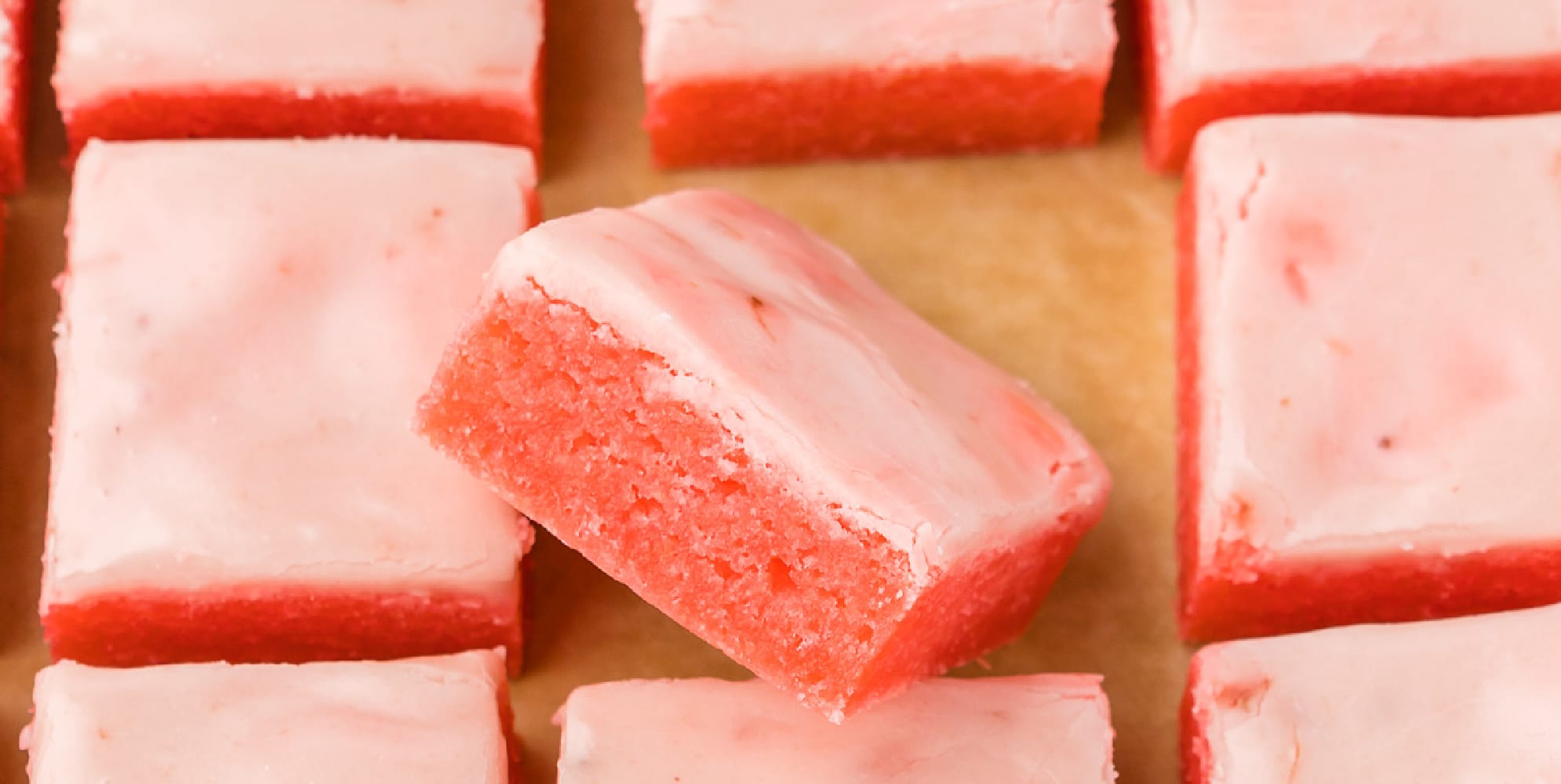 Strawberry brownies with glaze on parchment paper close up with one brownie turned to see the chewy edge and glaze better close up.