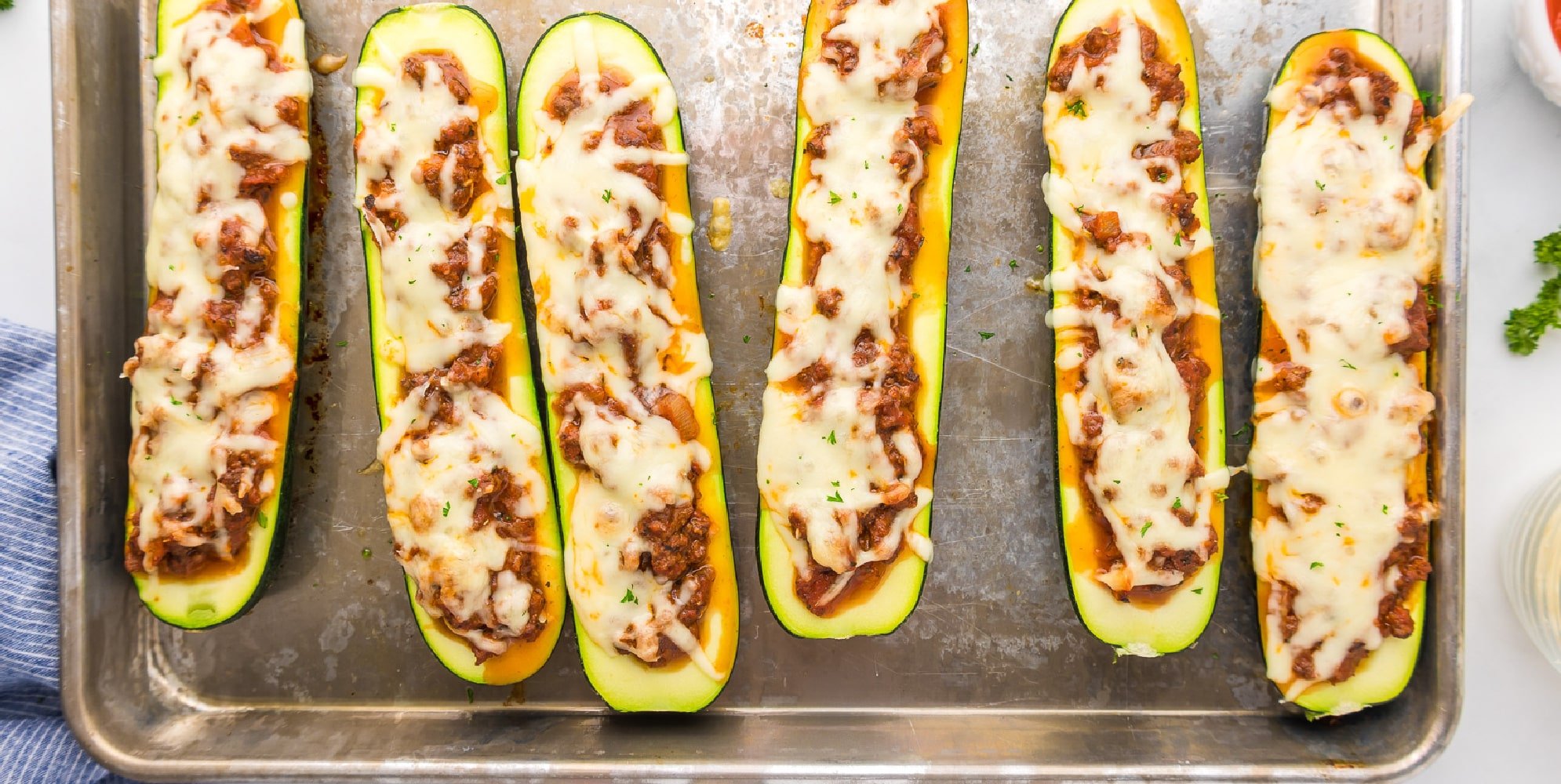 Six stuffed zucchini boats from overhead lined up on a sheetpan full of ground beef and cheese.