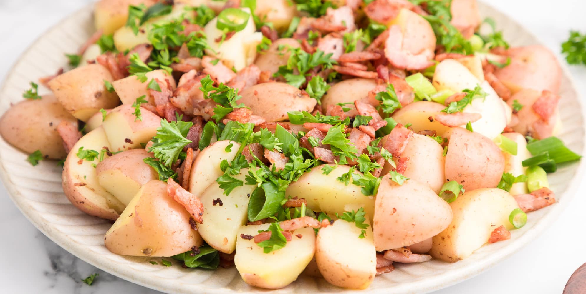 Wide view of warm german potato salad topped with bacon, parley and green onion pieces on a platter.