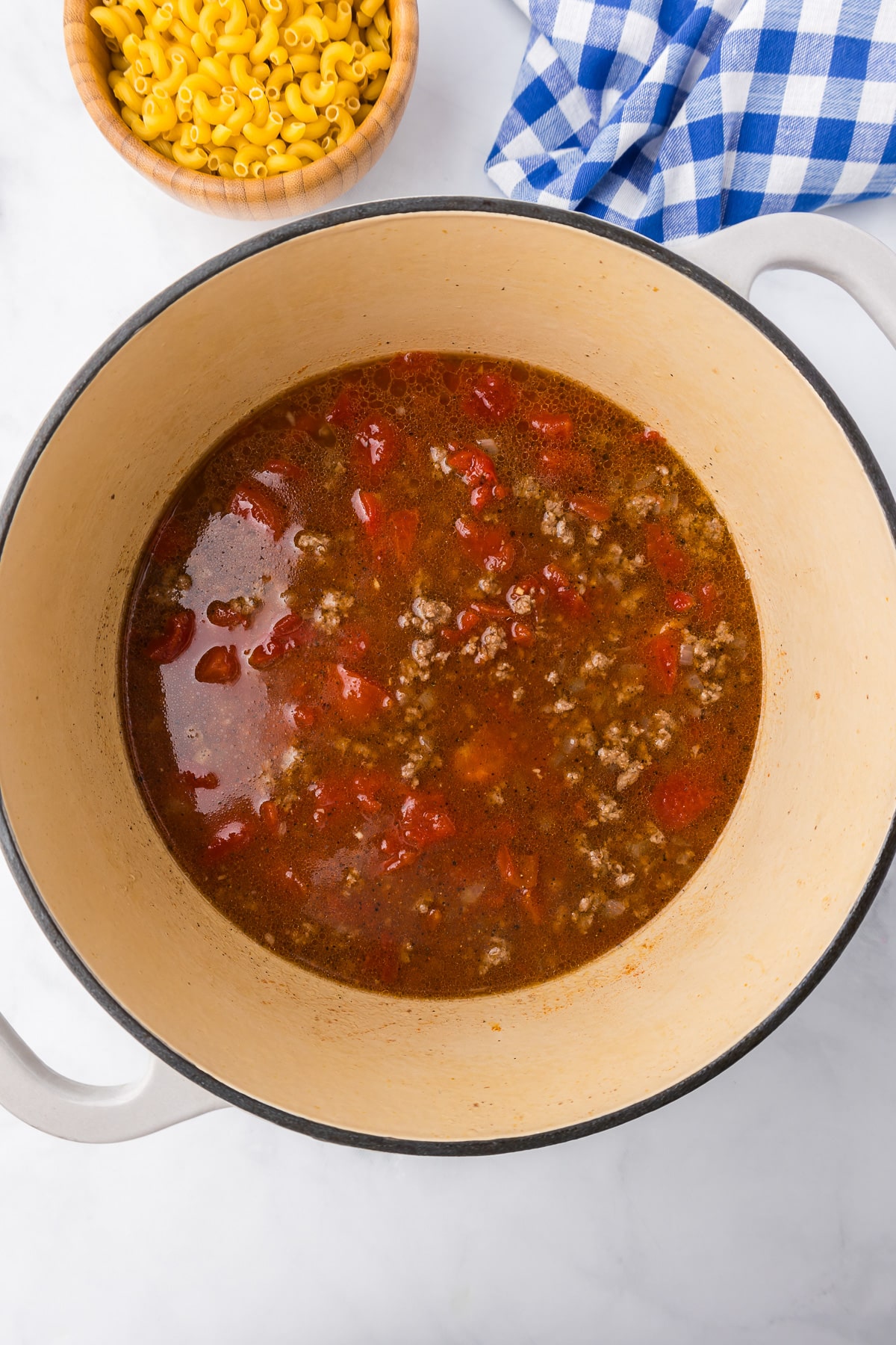Ground beef with broth and tomatoes in a large pot from overhead with a bowl of uncooked macaroni on the counter from nearby.