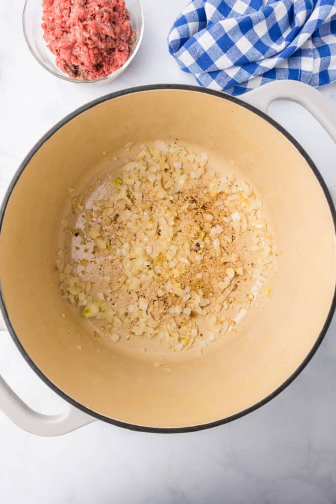 Diced onion being cooked in a pot from overhead with raw ground beef in a bowl on the counter nearby.