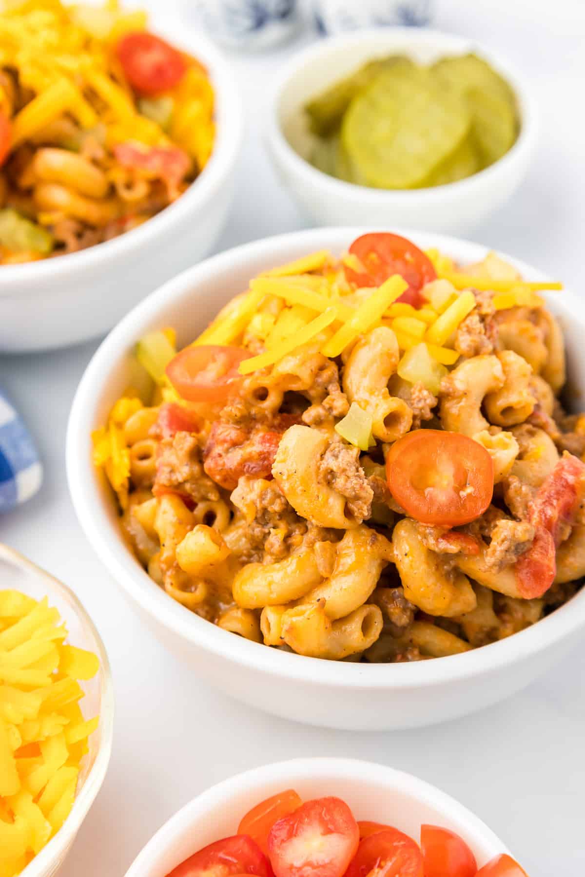Cheeseburger macaroni in a dinner bowl surrounded by more bowls of cheese, tomato and pickle toppings on the counter.