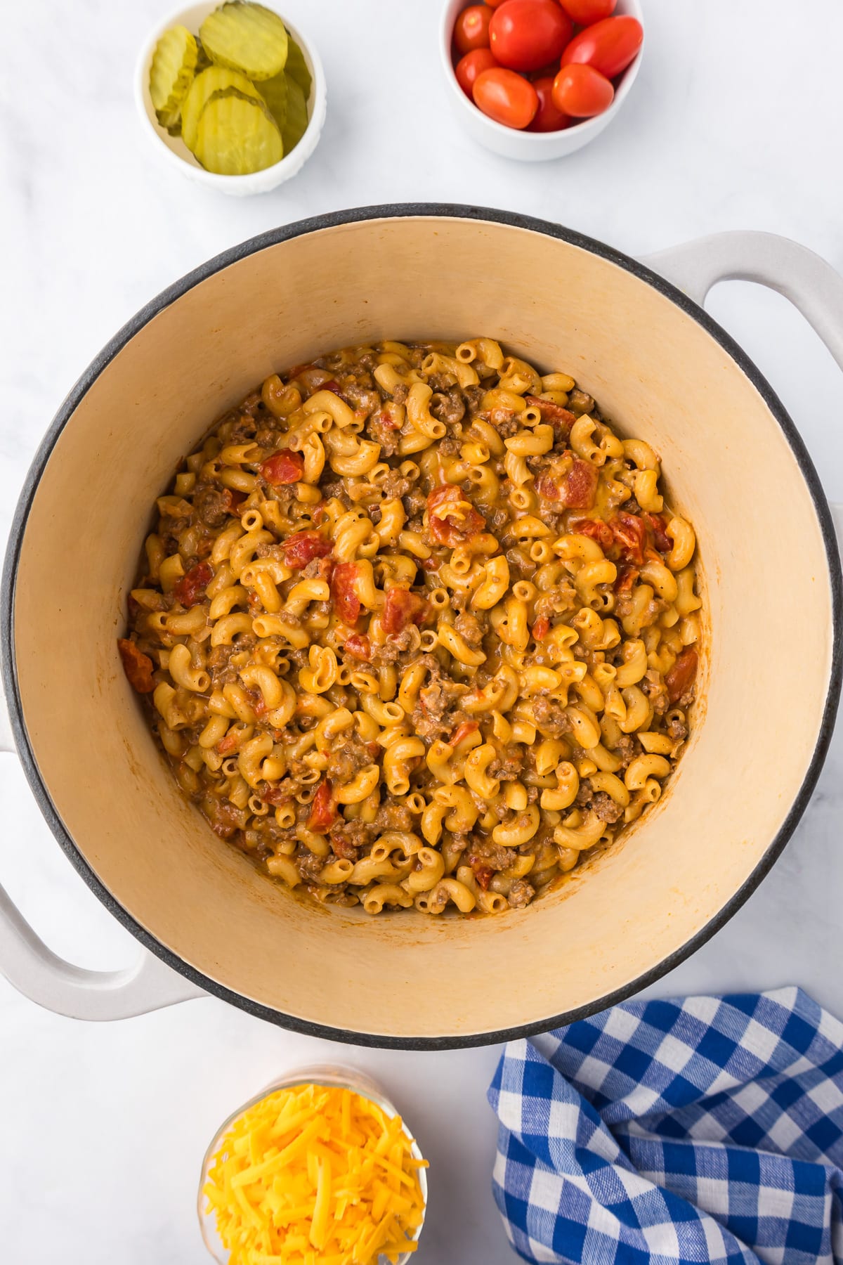 Cooked macaroni mixed with tomatoes and ground beef in a large pot from overhead with cheese, pickled and tomatoes in small bowls nearby.