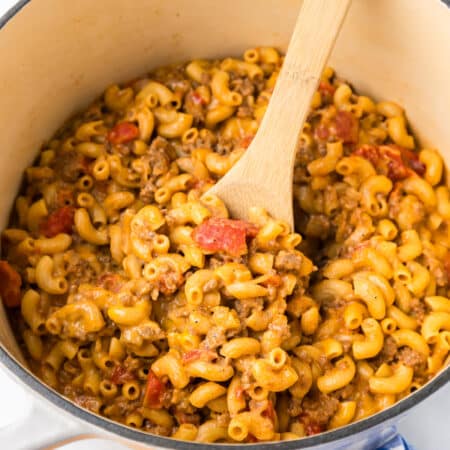 Close up inside a pot full of cheeseburger macaroni and pieces of tomato being stirred with a wooden spoon.