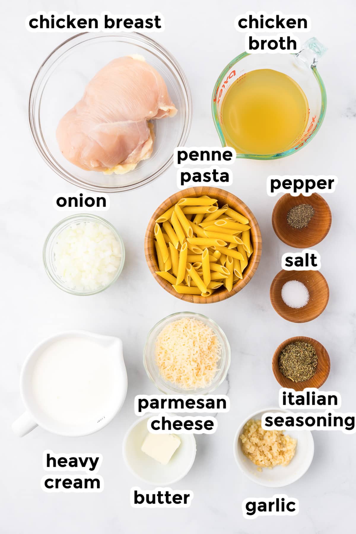 Ingredients for chicken penne alfredo in bowls on a counter from overhead with text labels.