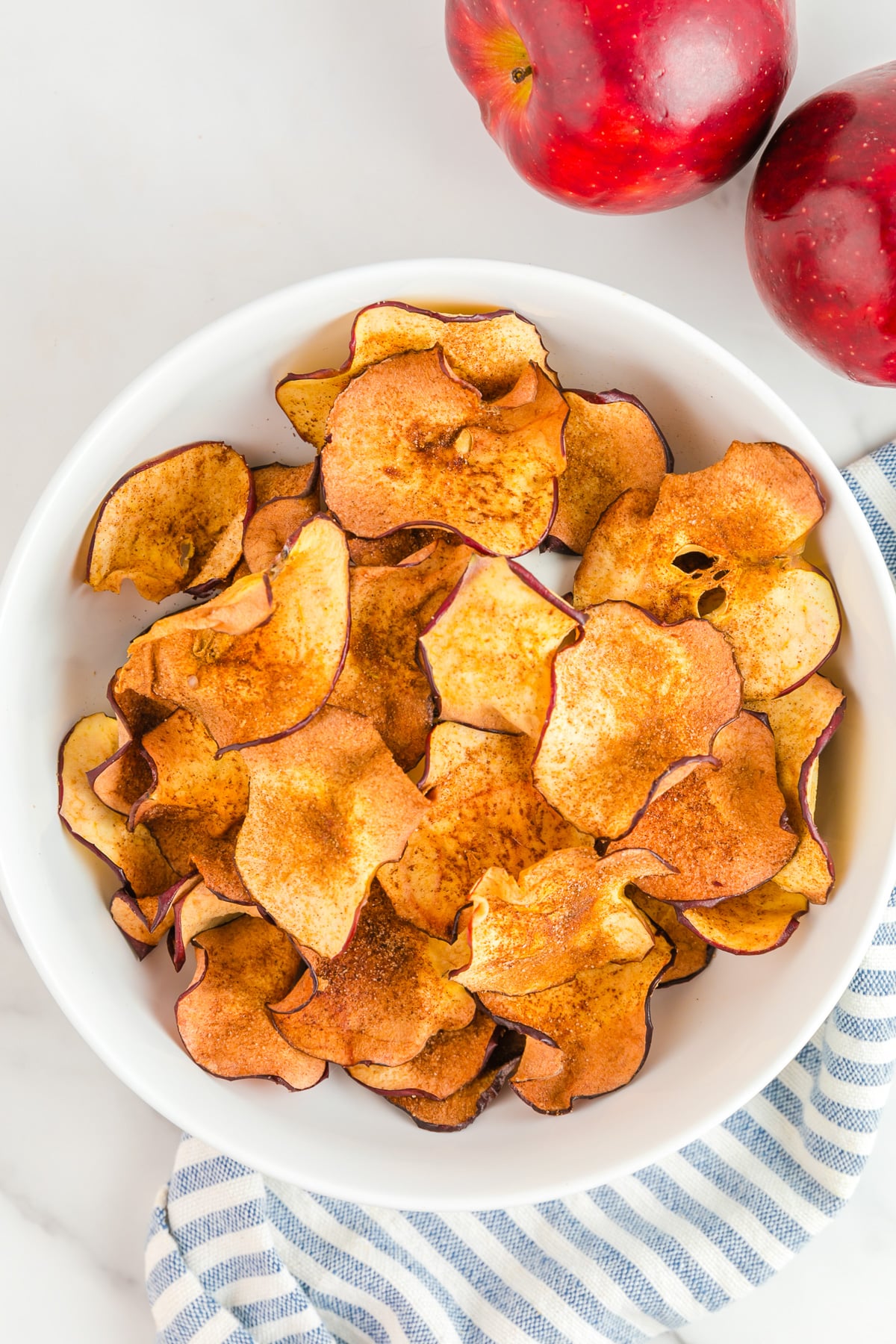 Apple chips in a bowl from above with two apples on the counter nearby.