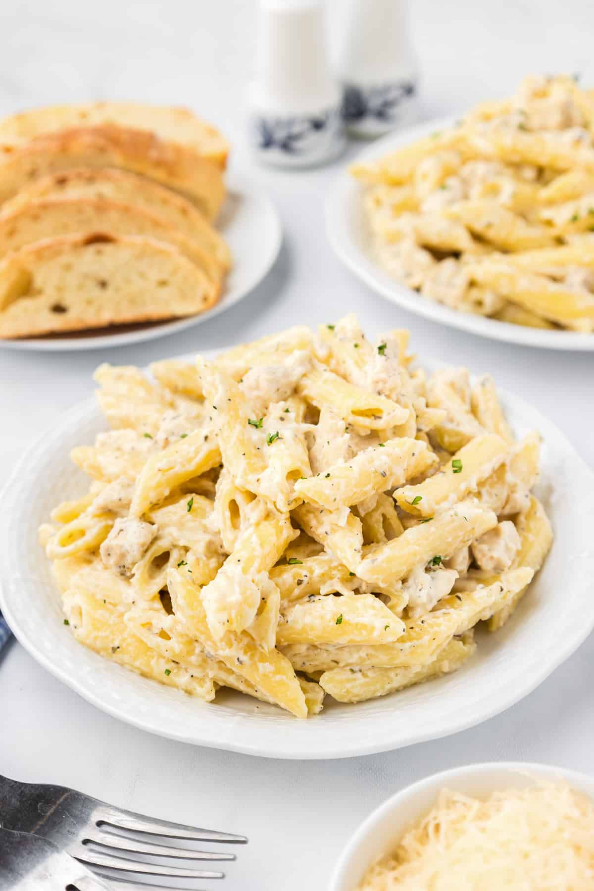 Chicken penne alfredo piled on a plate with bread and more pasta on plates in the background on the table.
