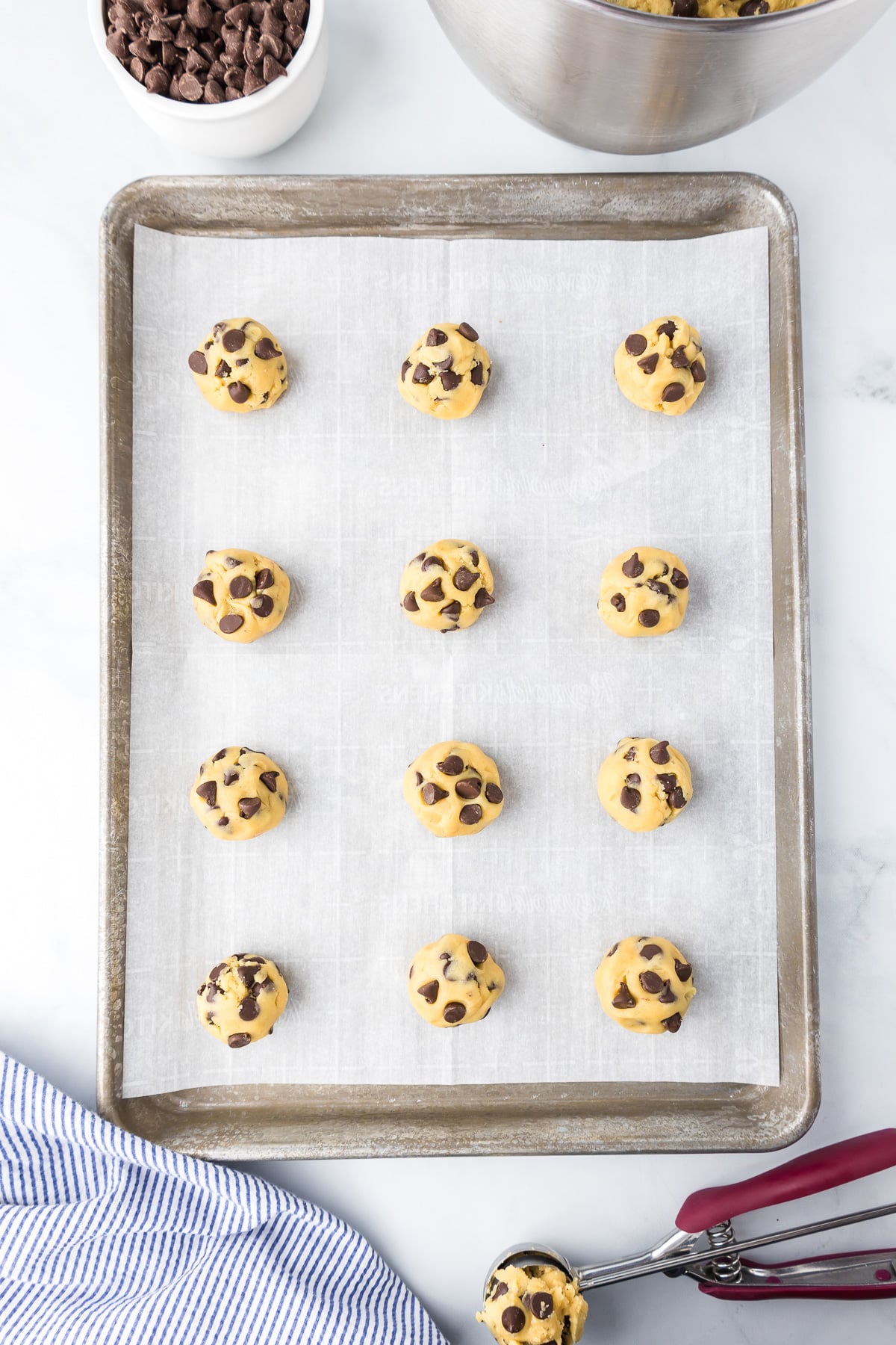 Chocolate chip cookie dough on a baking sheet line with parchment paper on the counter and a cookie scoop nearby.