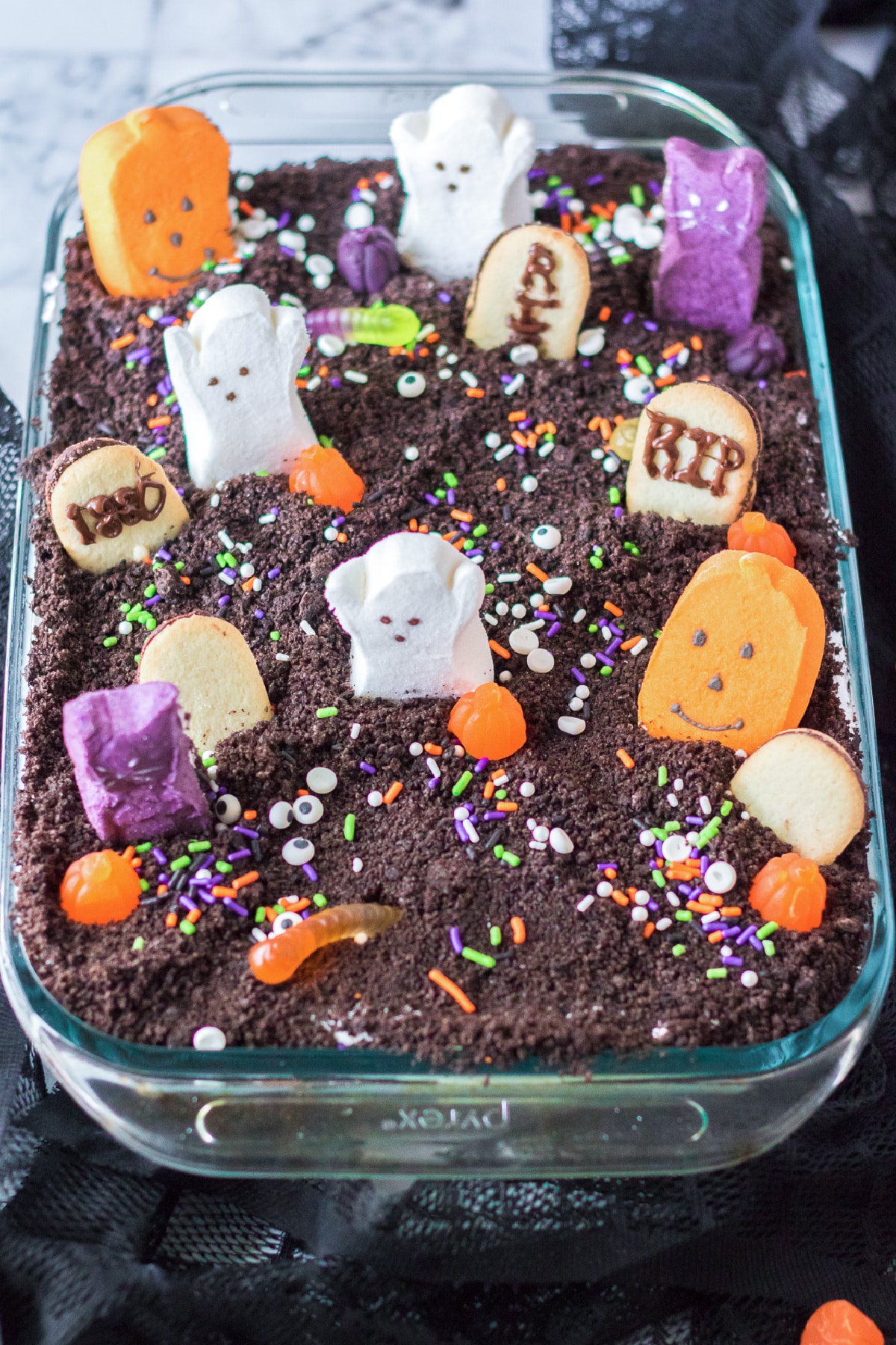 Chocolate graveyard Halloween poke cake from above covered in Halloween marshmallows, gummy words, cookie headstones, gummy words and Halloween sprinkles.