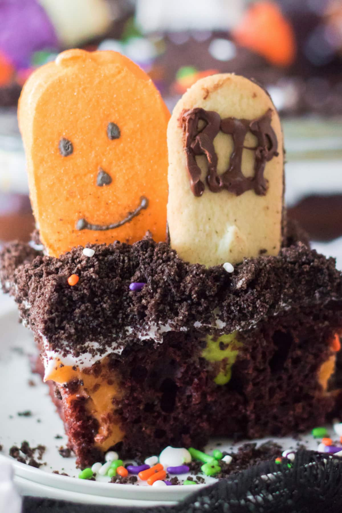 Chocolate poke cake topped with pumpkin marshmallow, cookie headstone and Halloween sprinkles sliced from the side on a plate.