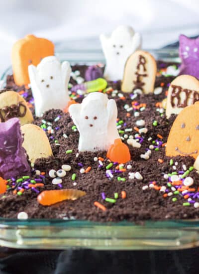 Square view of a chocolate ghost graveyard cake in a glass pan from the side decorated with Halloween marshmallows, gummy worms, and Halloween colored sprinkles.