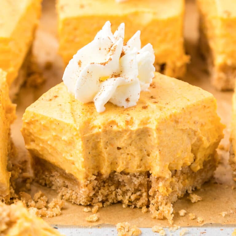 Close up square side view of a pumpkin cheesecake bar topped with a swirl of whipped cream on parchment paper with other slices of pumpkin bars in the background.