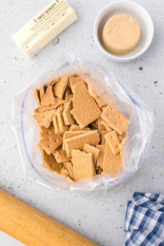 Graham crackers in a large zip-top bag open from overhead with brown sugar in a bowl and a stick of butter on the counter nearby.