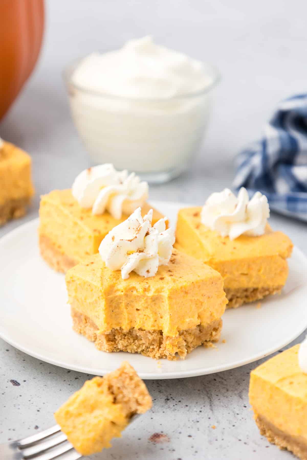 Three no bake pumpkin cheesecake bars on a plate from the side topped with whipped cream with the front bar missing a bite.