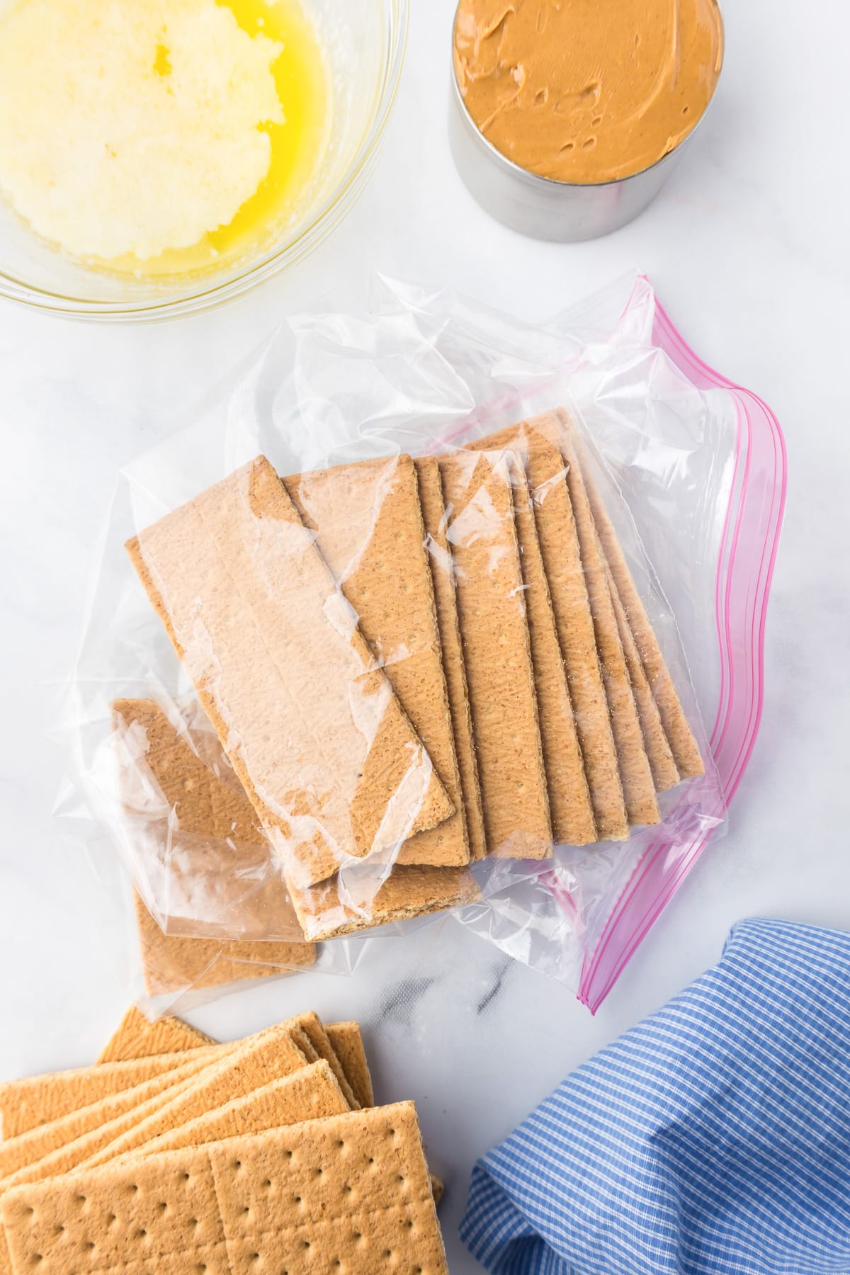 Graham crackers in a large ziplock bag from above with peanut butter and melted butter in bowls nearby on the counter from overhead.