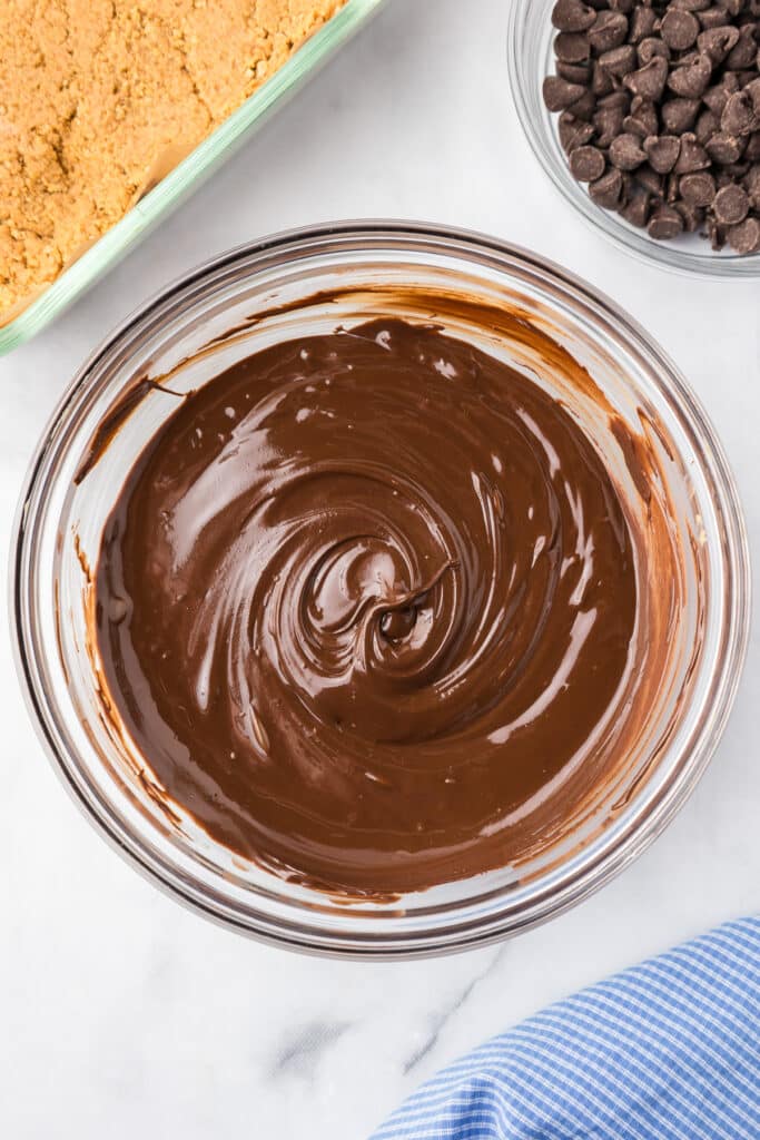 Chocolate melted in a bowl with the peanut butter graham cracker mixture in a pan nearby from overhead on a counter.