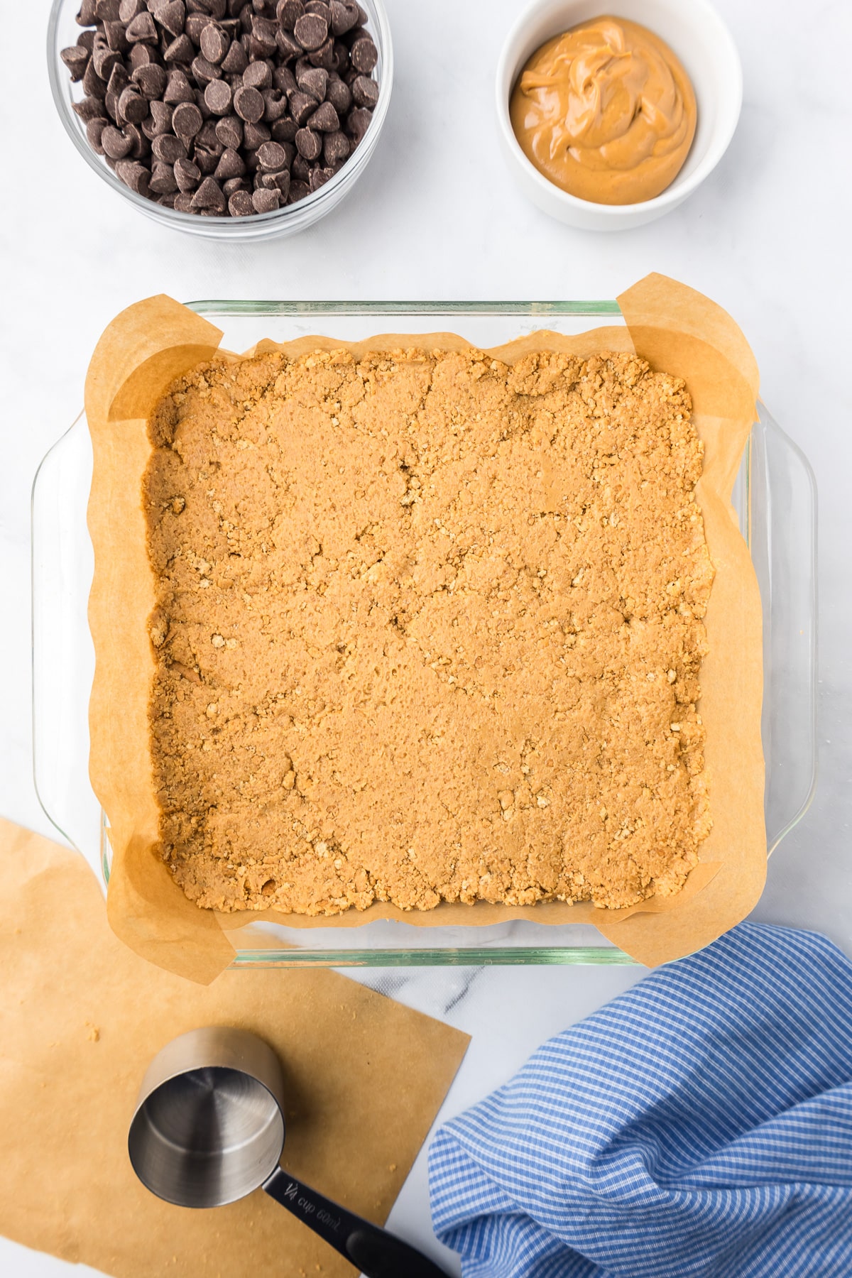 Peanut butter graham cracker mixture in a square pan from above with a measuring cup and parchment paper on the counter nearby.